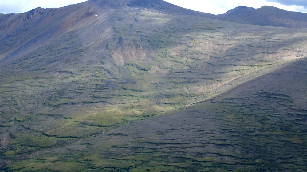 Landscape photograph showing vast hillsides covered with horizontal vegetation lines under a blue sky populated with fluffy clouds.