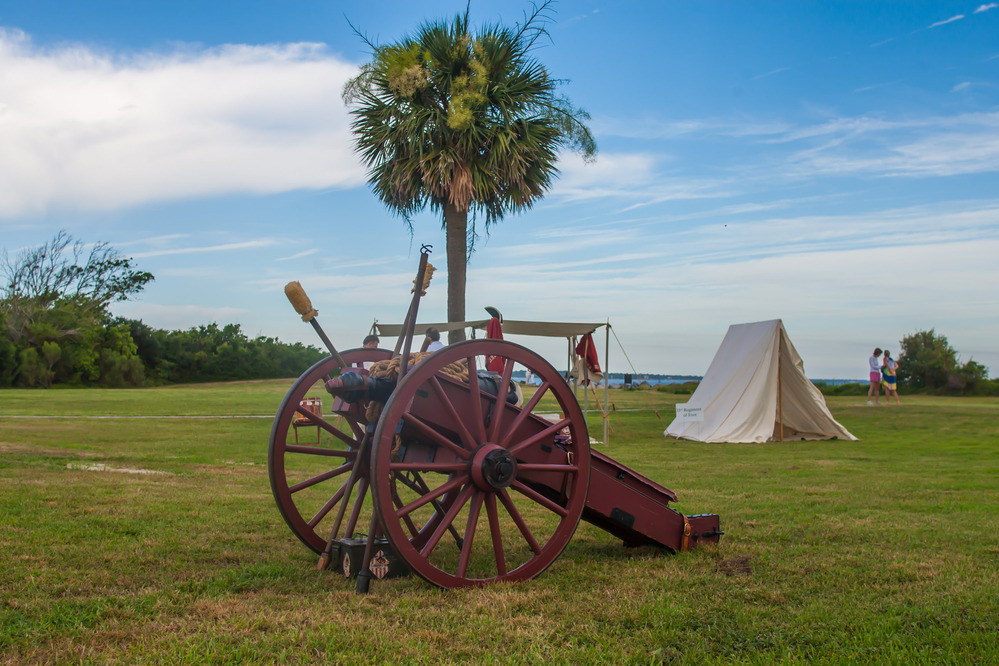 An 18th-century style military cannon on a reddish-brown wooden carriage sits in a grassy field, positioned in front of a lone palm tree. In the background, a white canvas A-frame tent stands near a thin line of trees under a bright blue sky with wispy clouds.
