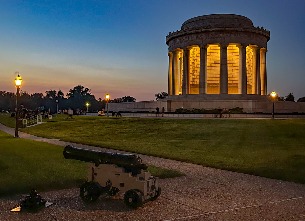 Large circular classical-style memorial building with prominent columns at night with a cannon and cannonballs in the forefront