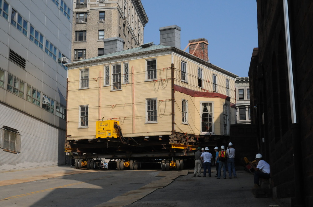 A yellow house on wheels on a street, very  close to the surrounding buildings.