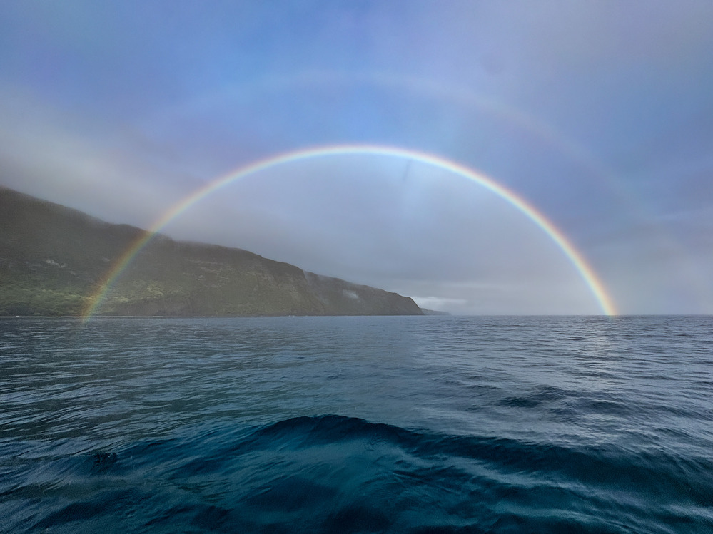 Outdoors; landscape view of almost flat blue water leading to cliff in background with full rainbow shining in middle of it all.