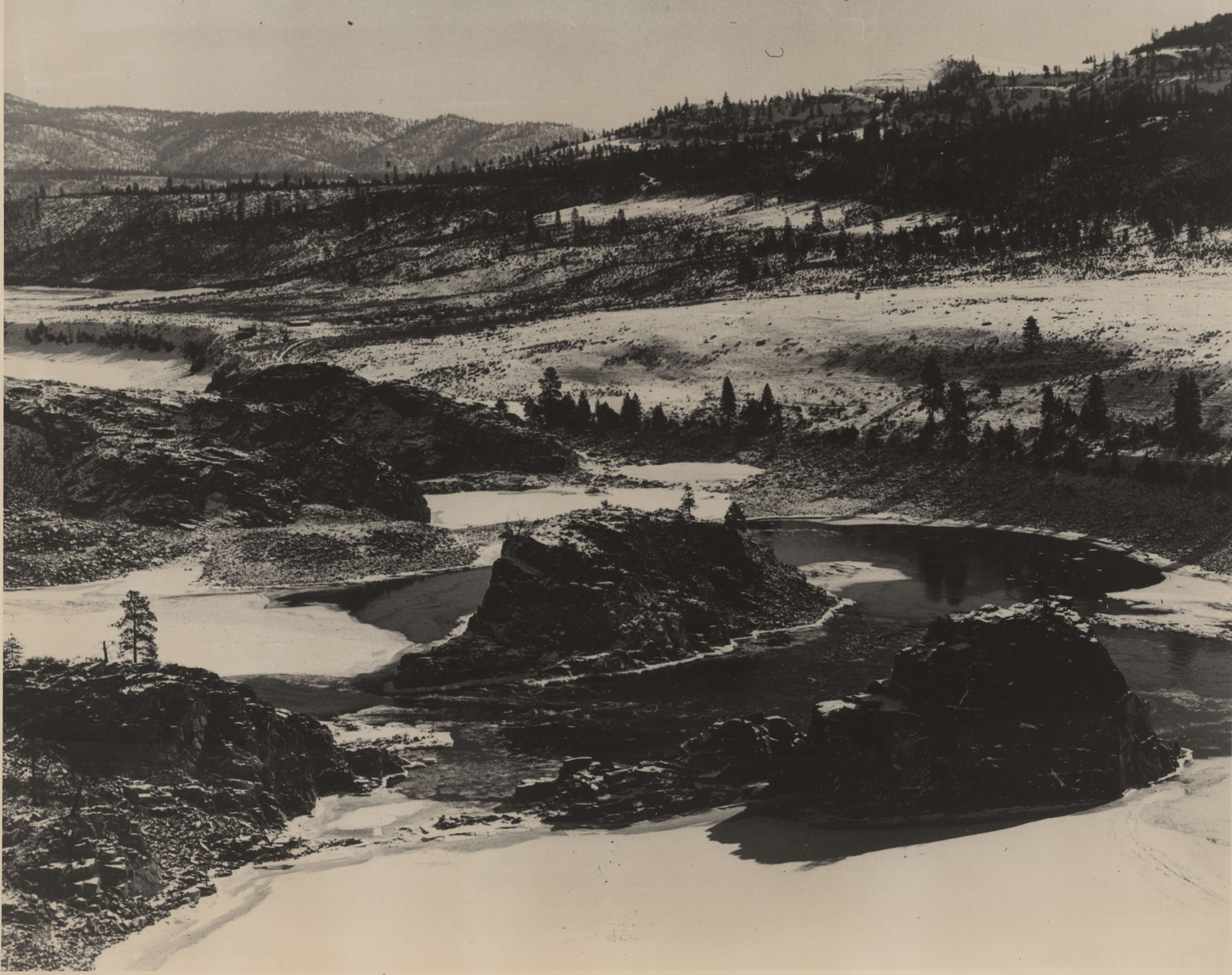 Black and white image of a partially frozen river meandering past large rock outcrops in a wide snowy, forested valley