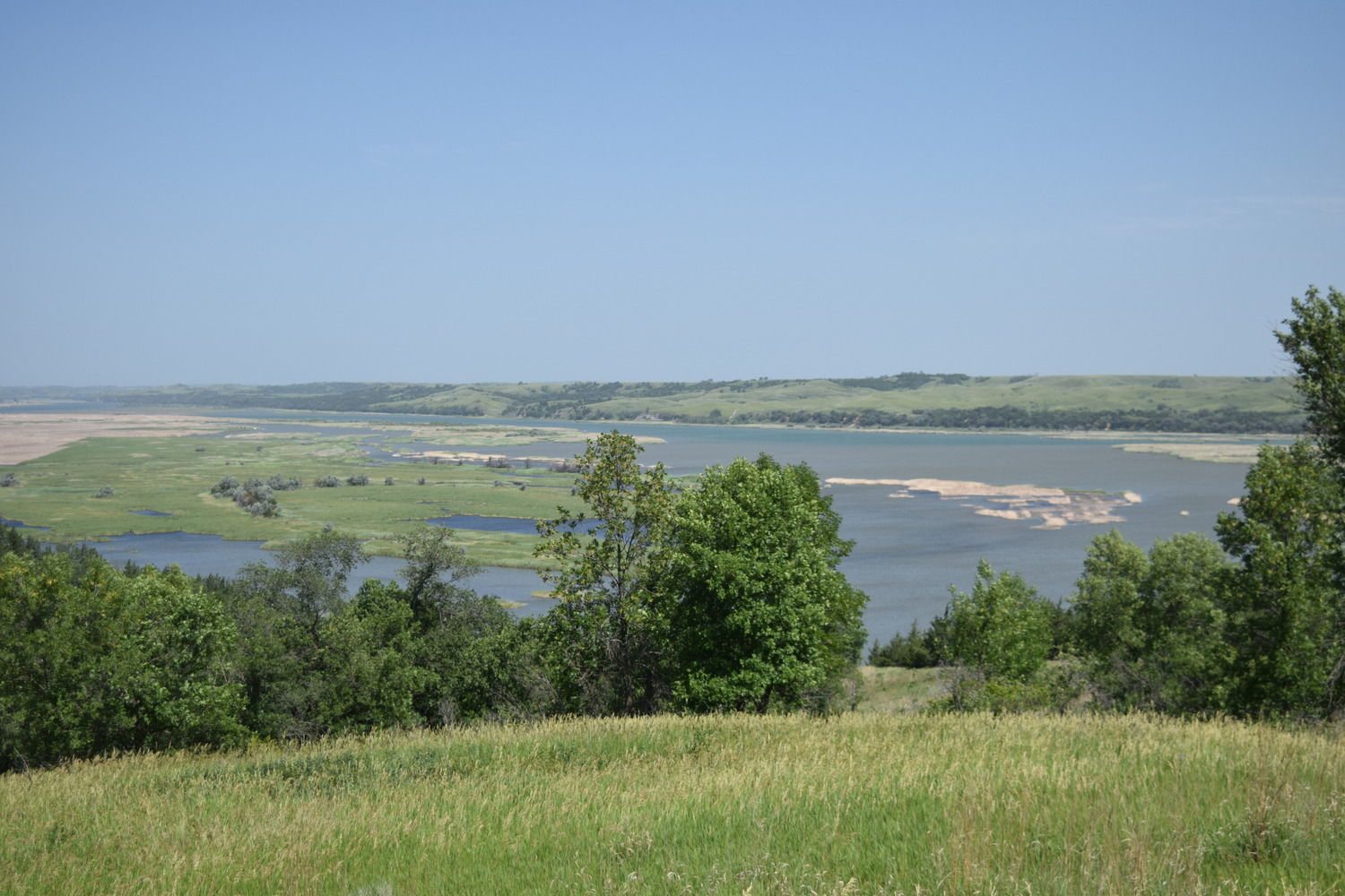 A wide, expansive river sits beneath a scenic overlook, with sandbars and islands. Grass and trees fill the foreground. 