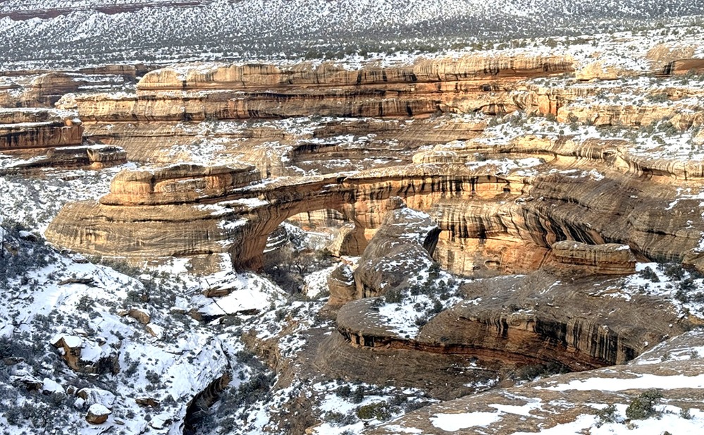 A stone bridge at a distance surrounded by snow-dusted shrubbery and standstone as streaks of sunlight paint the canyons.