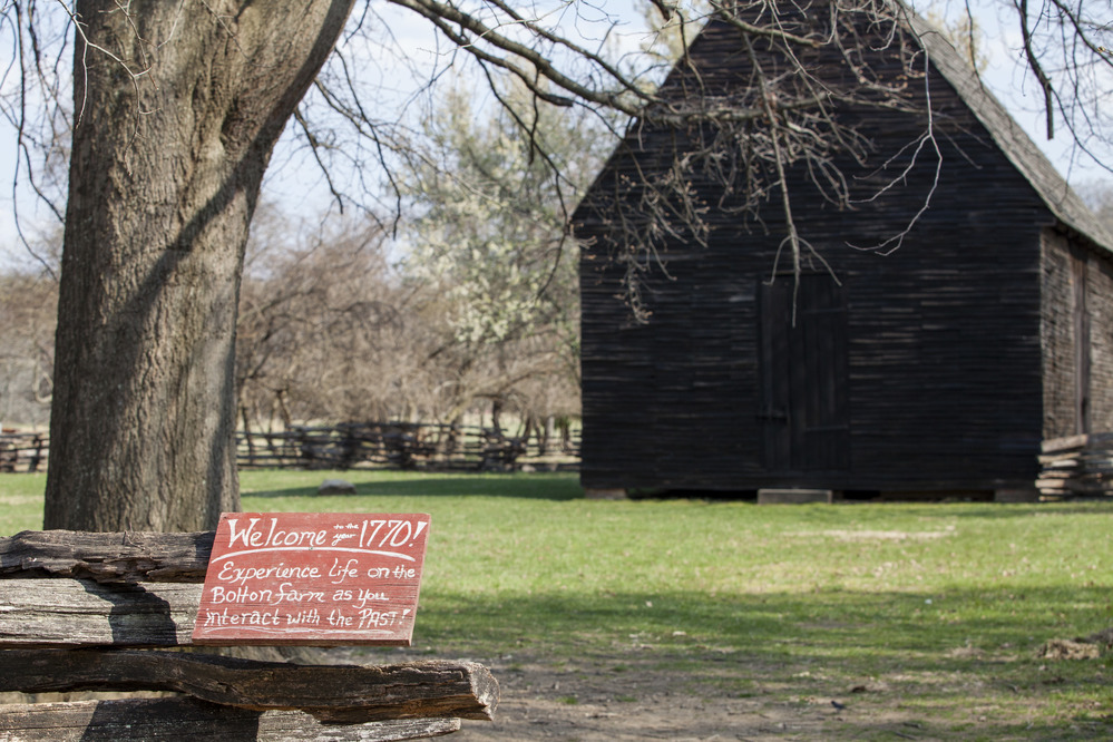 A small handwritten sign reads "Welcome to 1770! Experience life on the Bolton Farm as you interact with the past." It sits in front a one story farm building.