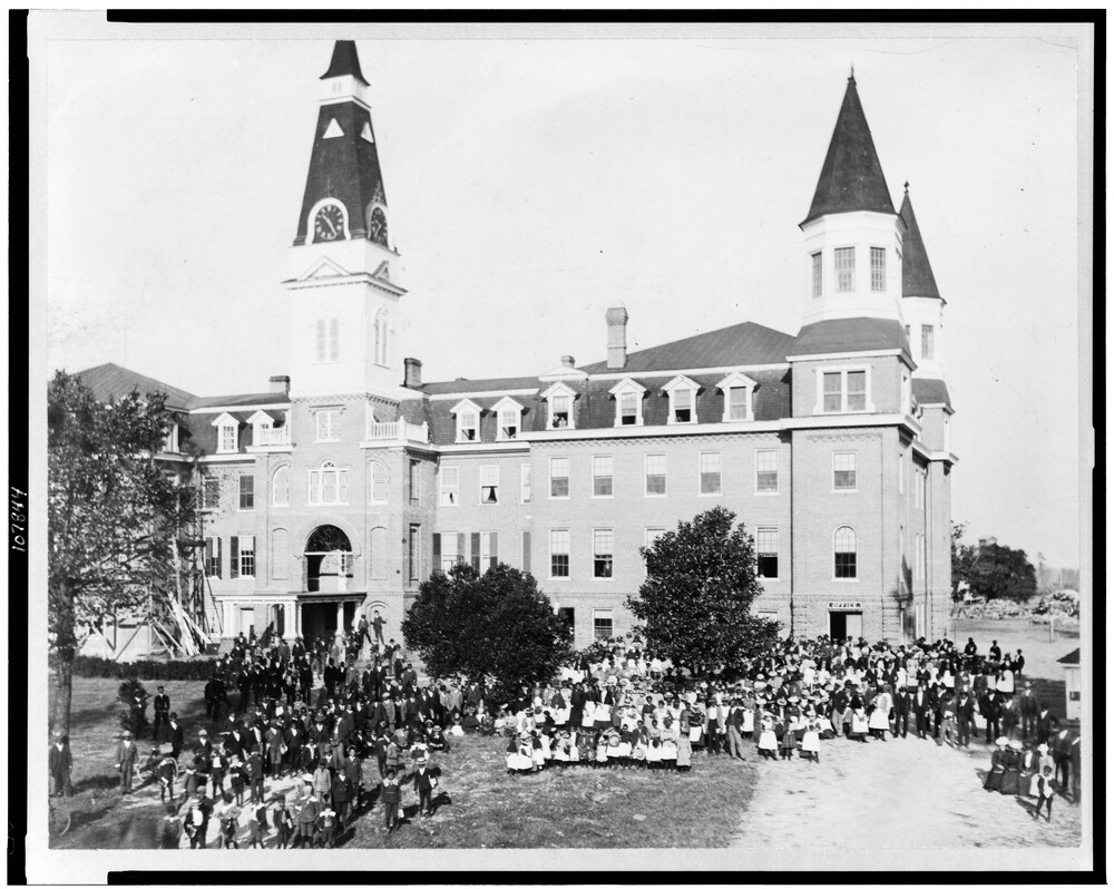 A group of students stand outside a large four-story building with three towers.