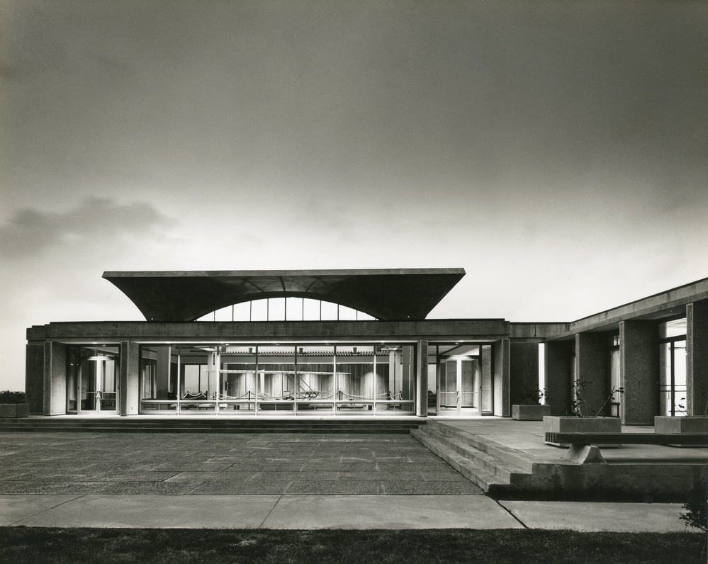 Black and white image of visitor center building with large open concrete space. Sky in background has wispy clouds.